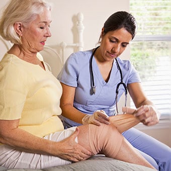 An at-home nurse caregiver wraps a woman's leg wound with a bandage.