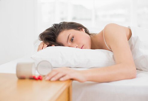 A woman half-awake lying on the bed next to a spilt bottle of pills on the nightstand. 