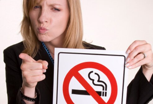 A woman points her finger and has a stern expression on her face while holding up a 'No Smoking' symbol.