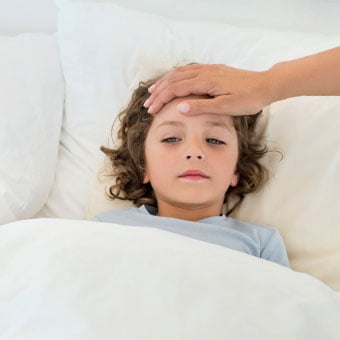 A boy sick in bed with an adult feeling his temperature by placing a hand on his forehead.